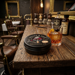 A round black tin of Stone & Silk Grooming Co. product sits on a rustic wooden bar counter in a barbershop-like setting, next to a filled cocktail glass and grooming bottles.