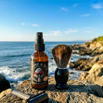 Amber spray bottle labeled Stone & Silky Grooming Co. pre shave oil beside a shaving brush and a razor on a rocky beach.