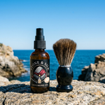 Amber spray bottle labeled Stone & Silky Grooming Co. pre shave oil beside a shaving brush on a rocky beach.