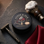 A round black tin of Stone & Silk Grooming Co. shaving soap sits on a dark slate surface, styled with a shaving brush, shaving razor, and red satin cloth.