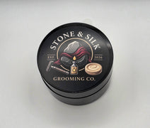 A round black metal tin of Stone & Silk Grooming Co. hair product, featuring a detailed label with a razor, oil bottle, and hair curl on a light gray background.