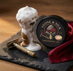 A round black tin labeled 'STONE & SILK GROOMING CO.' sits on a dark slate board beside a wooden shaving stump covered in lather and a brass-style safety razor.